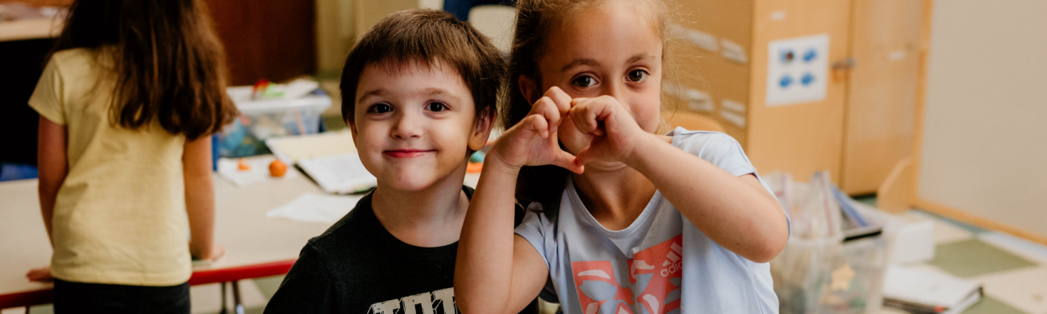 two students shown together in the classroom, one holds up her hands in the shape of a heart