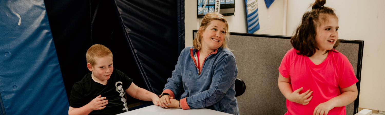 teacher seated at a table in classroom with a student and smiling at a student standing next to her