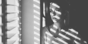 a young woman looks out of the window through blinds. photo is in black and white