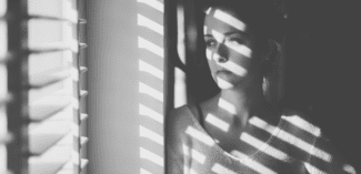 a young woman looks out of the window through blinds. photo is in black and white