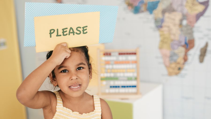 a young child is pictured holding up a sign that reads 'please' in a classroom