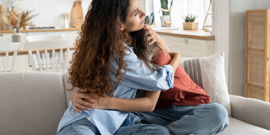 a parent is pictured hugging their child seated on a couch